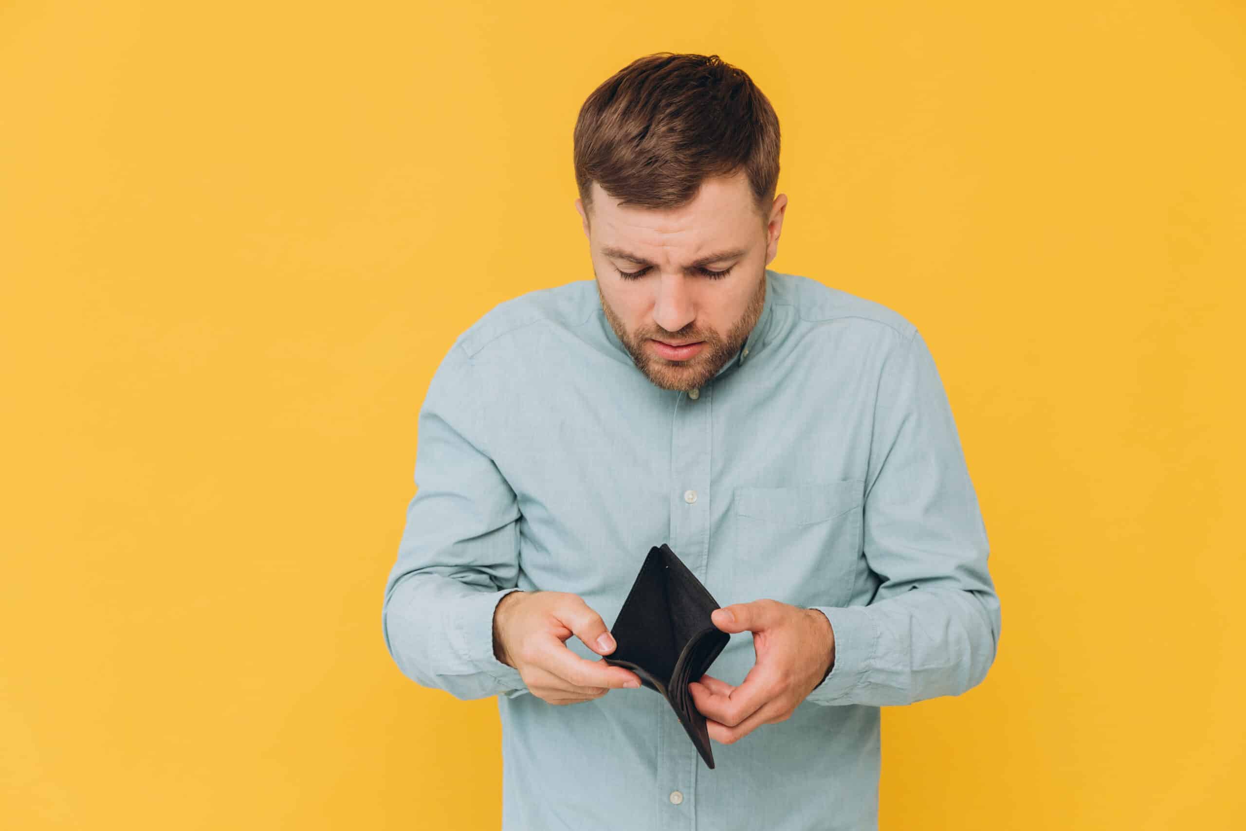 I'm Broke. Portrait of sad unshaven male bankrupt showing his empty wallet looking at camera. Upset man standing isolated over yellow studio background. Poverty Concept. Lack of money
