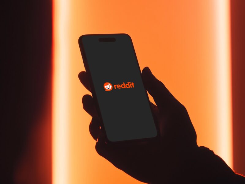 Silhouette of a person holding a smartphone displaying the Reddit logo on screen, against a red-orange light background.