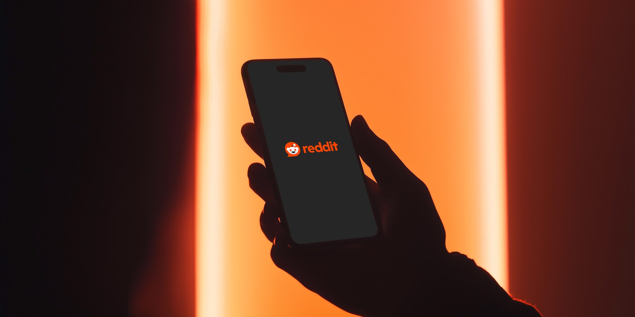Silhouette of a person holding a smartphone displaying the Reddit logo on screen, against a red-orange light background.