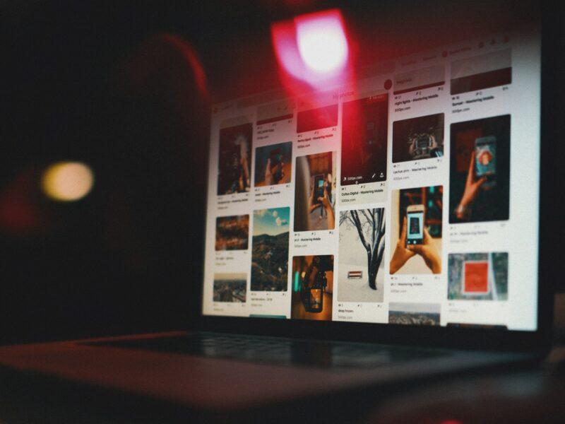 A laptop screen glowing in a dark room, displaying a Pinterest gallery of various photos and pins, with a soft red light reflecting across the top of the screen.