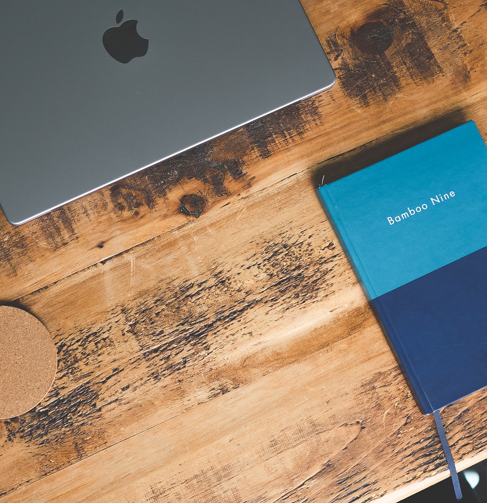 Close-up of a wooden desk with a silver MacBook, a blue Bamboo Nine notebook, and a cork coaster — modern workspace setup at Bamboo Nine digital marketing agency.