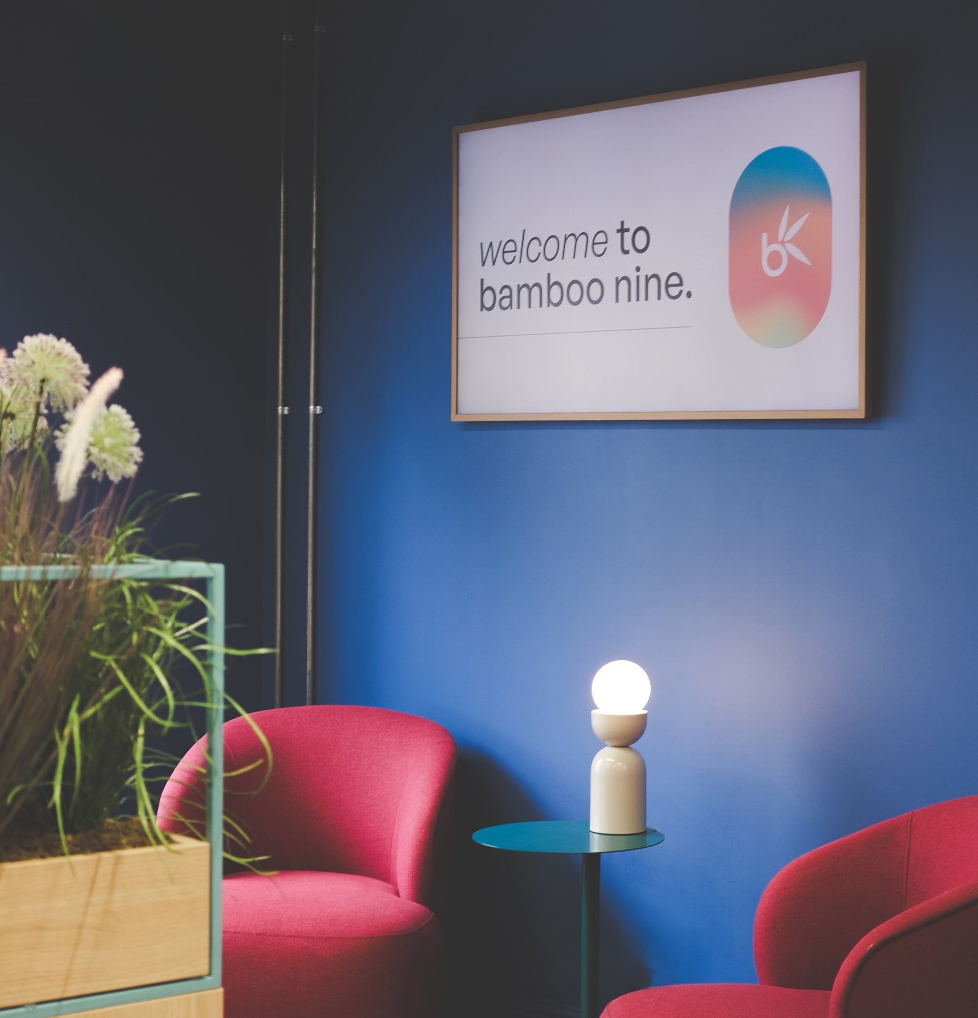 Bamboo Nine office reception area with blue walls, red chairs, modern lighting, indoor plants, and a wall sign that reads ‘Welcome to Bamboo Nine’.