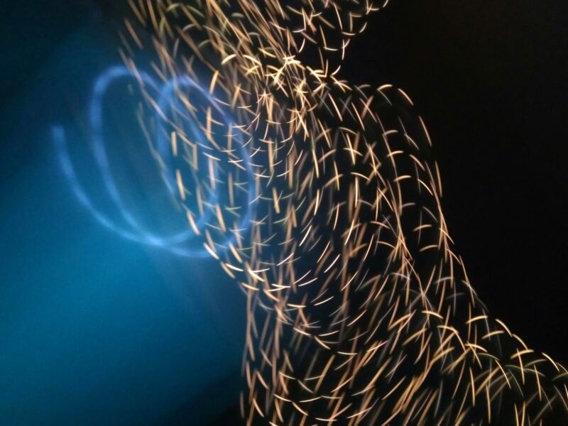 Abstract light trails forming a flowing, wave-like pattern of golden sparks and soft blue glow against a dark background.