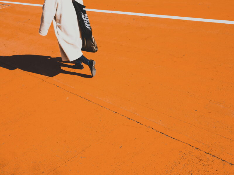 Person in a long beige coat walking across a bright orange pavement, casting a strong shadow, with a black tote bag over their shoulder and a white line marking the ground in the background.
