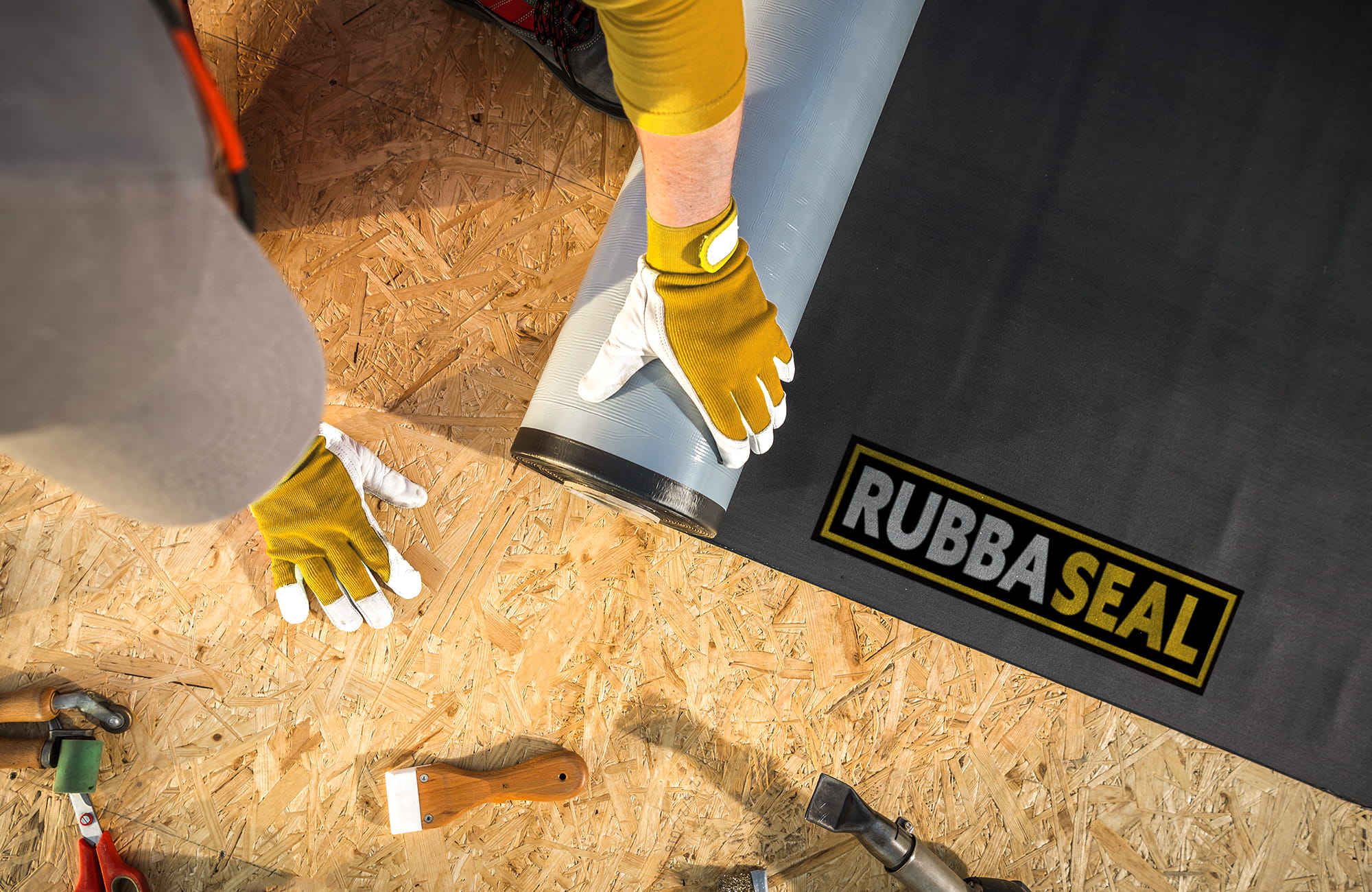 Overhead view of a person wearing yellow gloves rolling out a grey rubber membrane onto a wooden surface, with installation tools nearby and a “Rubba Seal” branded sheet visible.